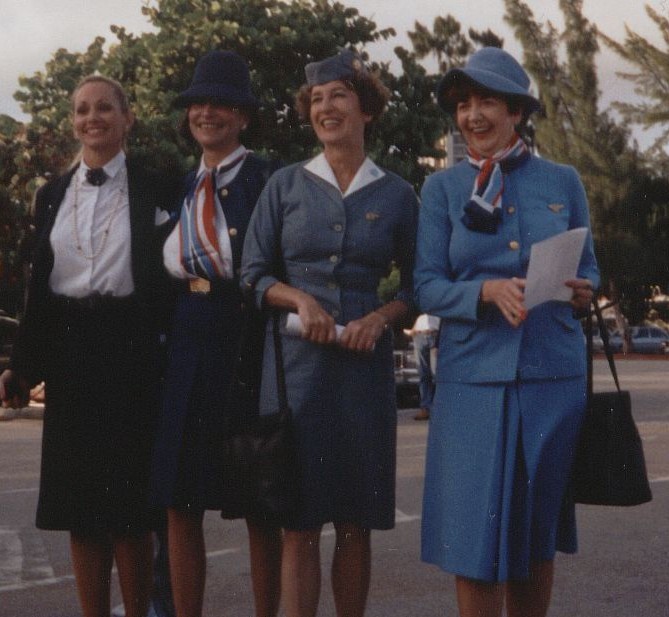 1991 Three  generations of Pan Am flight attendant uniforms.  On the left is Pan Am's final uniform.  Originally designed by Adolfo in the 1980s and later modified to include the round neck tie pictured.  The 1970s Edith Head uniform with signature red, white and blue scarf is worn by the flight attendant on the right (in Pan Am blue) and second from left (Navy Blue).  The early 1960s uniform designed by Don Loper is seen on the flight attendant second from the right.
